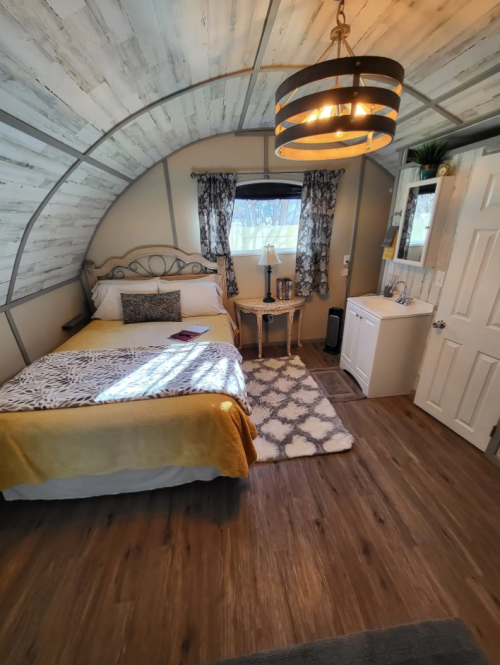 Cozy bedroom with a curved ceiling, a bed, nightstand, and decorative window curtains. Warm lighting and wooden floors.